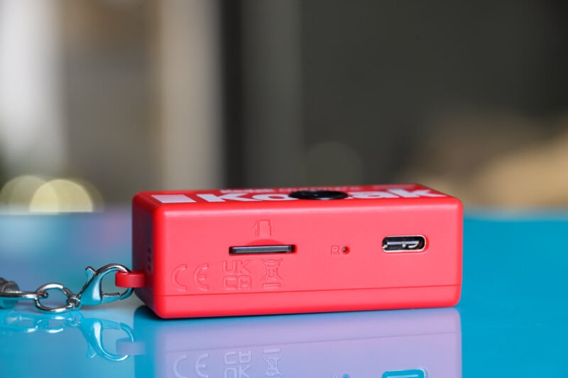 Primer plano de un pequeño dispositivo Kodak rojo con llavero, equipado con ranura para tarjeta microSD, puerto USB-C y marca de certificación, colocado sobre una superficie azul brillante.