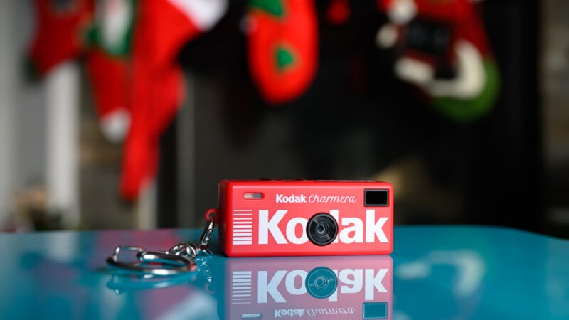 Una pequeña cámara de llavero Kodak roja se encuentra sobre una superficie azul, y medias navideñas borrosas cuelgan al fondo.