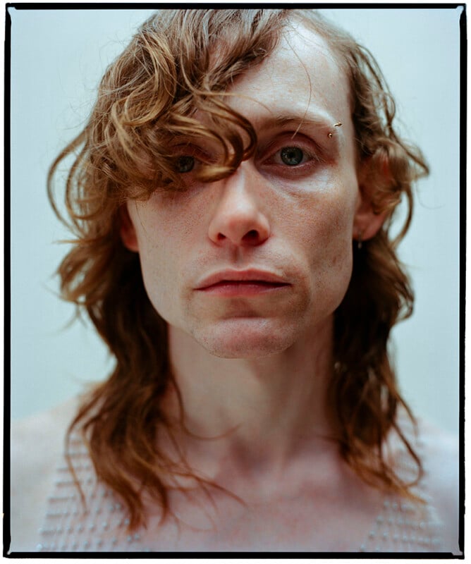 Un hombre de piel clara y cabello castaño ondulado hasta los hombros mira directamente a la cámara. Un ojo está parcialmente cubierto de pelo. Tienen expresiones suaves y usan blusas texturizadas sin mangas. El fondo es neutro y sencillo.