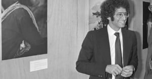 A man in a suit and tie with curly hair and glasses smiles while standing indoors next to a wall displaying black-and-white photographs.