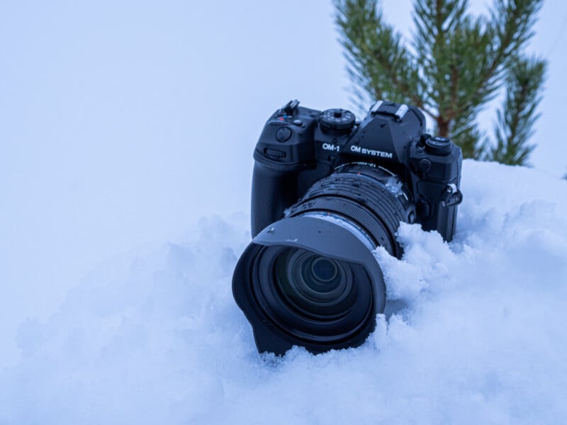 A black OM SYSTEM camera with a large lens is partially buried in snow, with a small pine tree visible in the background.