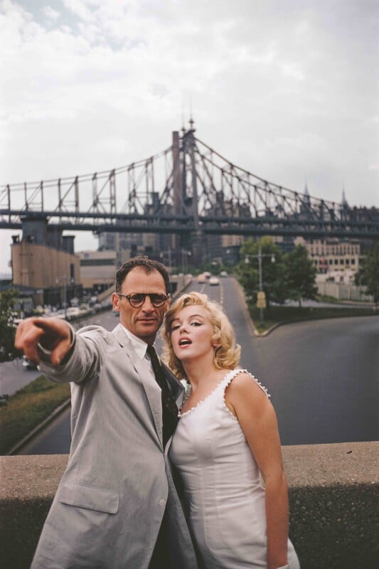 A man in a suit and glasses points forward while standing next to a woman in a white dress. They pose in front of a large steel bridge on a cloudy day, with a city street and trees visible below.
