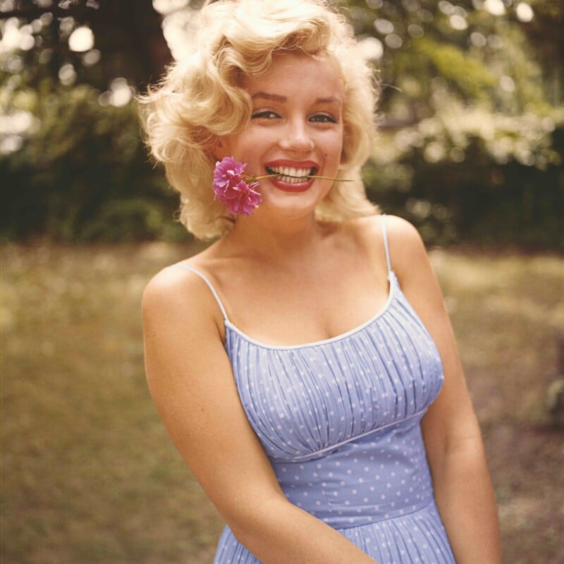 Smiling woman with blonde hair in a light blue, polka-dot dress stands outdoors, holding a pink flower in her mouth. The background is blurred greenery, creating a warm and cheerful atmosphere.