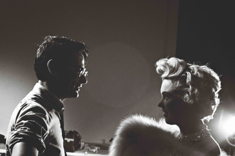 A black-and-white photo showing a man in glasses facing a woman with curled blonde hair and a fur wrap, softly lit, as they appear to be talking on a film or photo set.