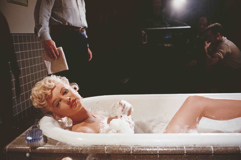 A woman with short blonde hair and red lipstick reclines in a bubble-filled bathtub, gazing upward. People and studio lights are visible in the background, suggesting a film or photo shoot set.