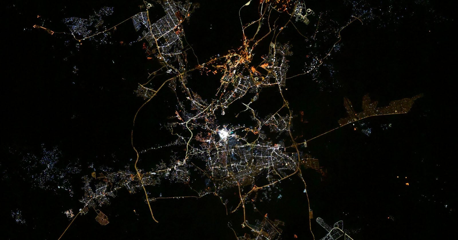 A city is illuminated at night, with bright lights outlining roads and neighborhoods, seen from above against a dark background. The city center appears brightest, with networks of lights branching outward.