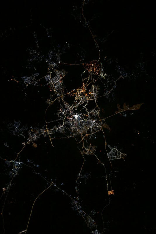 A nighttime aerial view of a city with a network of illuminated streets and highways forming bright, branching patterns against a dark background. Dense clusters of lights indicate urban centers and surrounding areas.