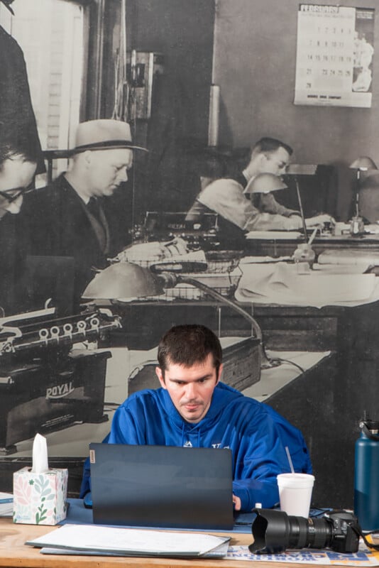 A man in a blue hoodie works intently on a laptop at a desk cluttered with a drink, tissue box, camera, and water bottle. Behind him is a large black-and-white mural of office workers at desks with typewriters.