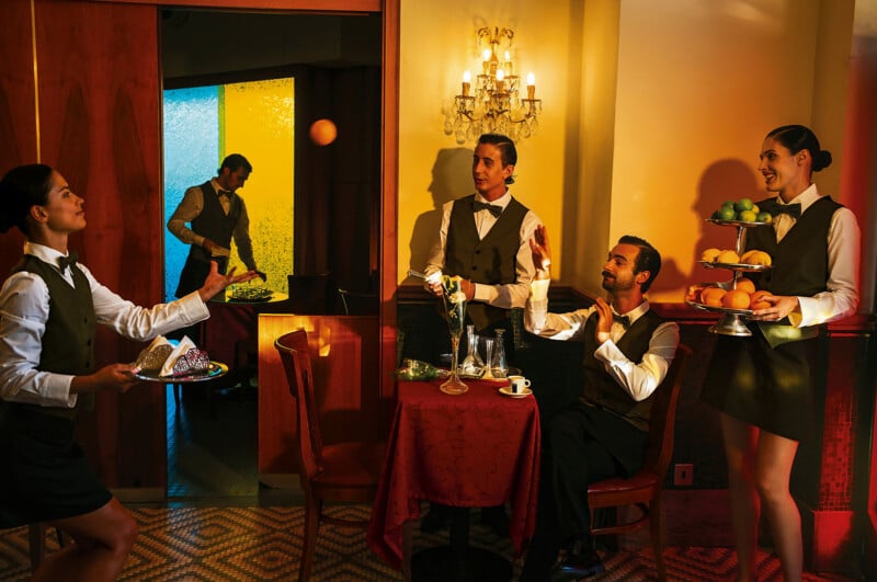 Four servers in uniforms are in a restaurant; one serves fruit, another juggles an orange, a third presents dessert, and a seated server gestures with a hand. A server in the background is arranging food near a counter.