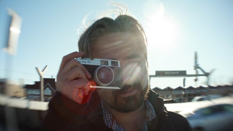 Un hombre con cabello castaño claro y barba sostiene una cámara antigua frente a su rostro al aire libre, con la luz del sol y el destello de la lente oscureciendo parcialmente su rostro. En el fondo borroso se pueden ver señales de tráfico y edificios.