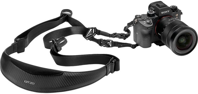 A Sony digital camera with a lens attached is connected to a black Gitzo-branded camera strap with padded shoulder support, lying on a white background.