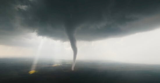 A dark, swirling tornado extends from a stormy sky to the ground over a flat landscape, with beams of sunlight breaking through the clouds in the background.