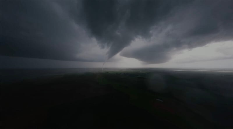 Un tornado oscuro con forma de embudo aterrizó desde lejos bajo siniestras nubes de tormenta sobre un paisaje rural llano, con parches de campos verdes visibles debajo.