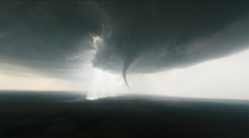 Un tornado oscuro desciende de las nubes de tormenta a una vasta tierra plana, con la luz del sol atravesando las nubes al fondo.