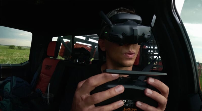 La persona sentada en el coche lleva gafas de realidad virtual y se concentra en manejar el dispositivo de control remoto. La luz del sol se filtra a través de las ventanas, revelando los campos exteriores y el equipo en el asiento trasero.