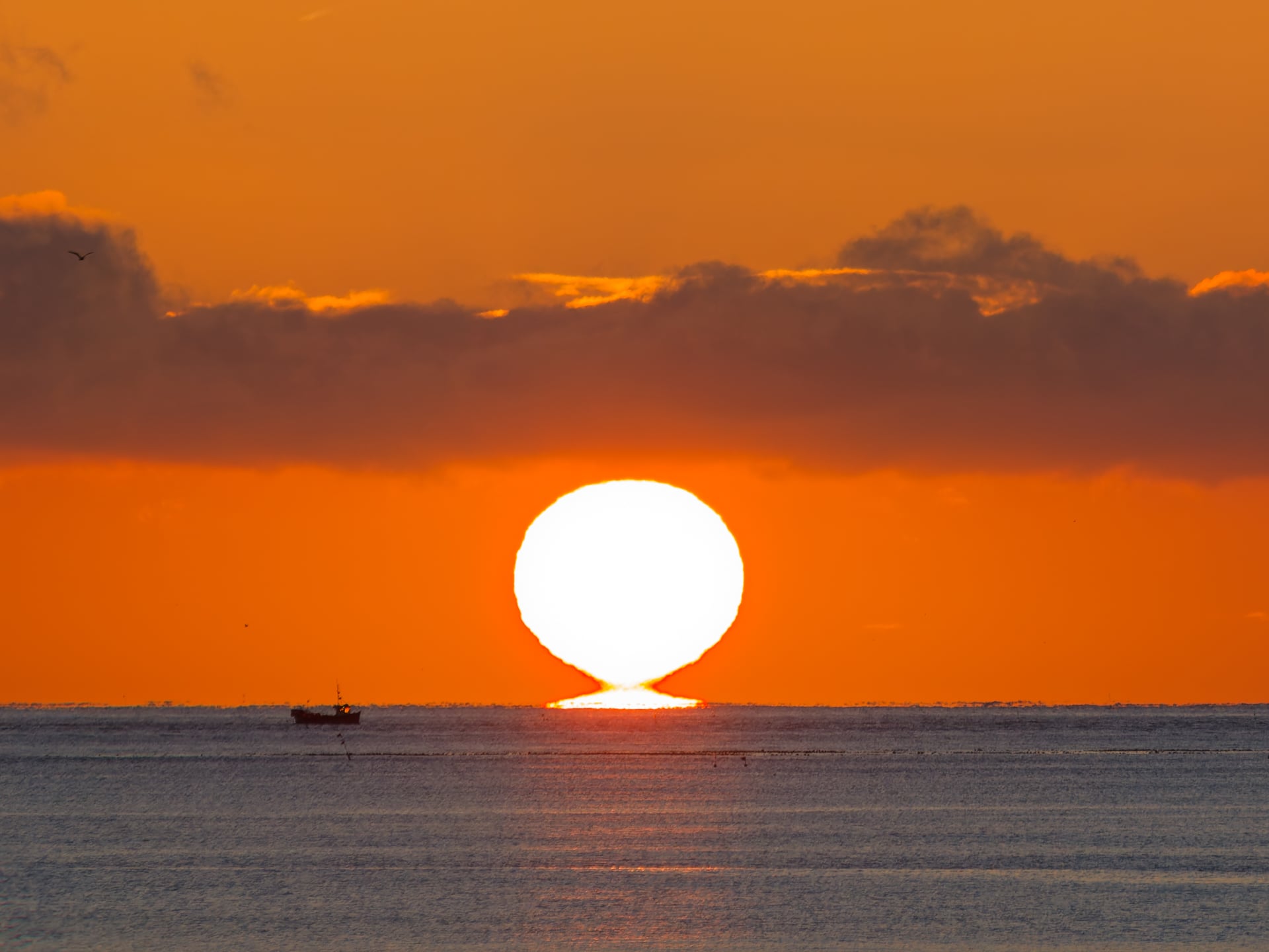 Un enorme sol de color naranja brillante sale y se pone sobre el océano, tocando el horizonte. Un pequeño barco flota en las tranquilas aguas a la izquierda del sol. El cielo estará mayormente despejado con nubes oscuras sobre el sol.