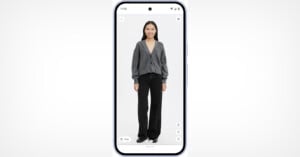 A smartphone displays a smiling woman wearing a gray cardigan and black pants, standing against a plain white background.