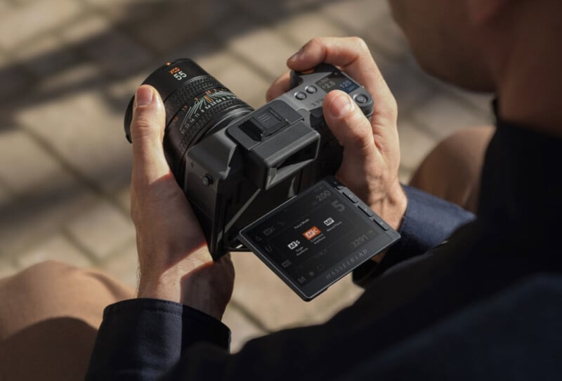 A person holds a digital camera with a large lens, tilting the touchscreen display to review camera settings or images. The camera is being operated outdoors, and the person's hands are clearly visible.