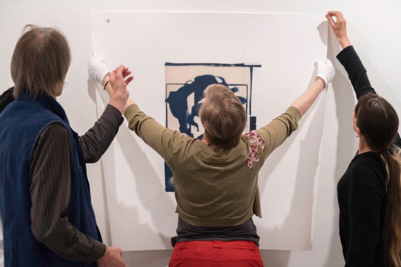 Three people hang a large piece of artwork on a white wall. The person in the middle wears gloves and holds the bottom, while the others adjust the top corners. The artwork features a blue abstract design.