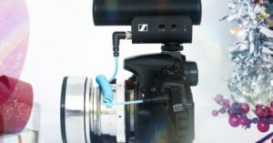 A black DSLR camera with a large lens and an external microphone attached sits among festive decorations, including silver tinsel, red berries, and a wrapped gift, against a white background.