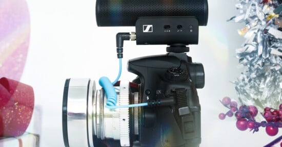 A black DSLR camera with a large lens and an external microphone attached sits among festive decorations, including silver tinsel, red berries, and a wrapped gift, against a white background.