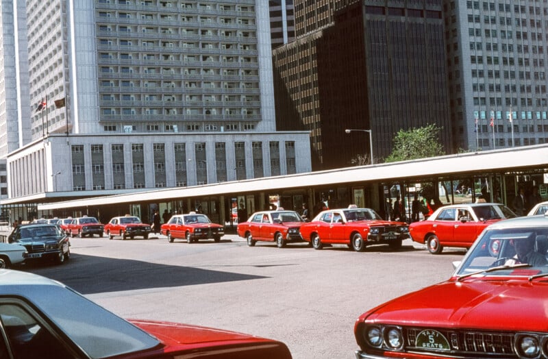 Una fila de taxis rojos espera en una parada de taxis frente a altos edificios de oficinas de la ciudad; algunos conductores son visibles en el interior.