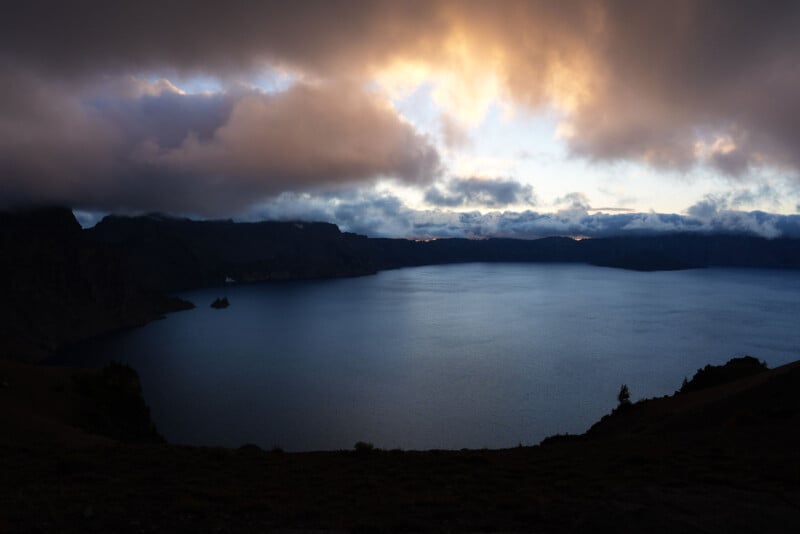 Al atardecer, un amplio lago está rodeado de montañas bajo un cielo nublado. El agua está tranquila y oscura, mientras las nubes adquieren tonalidades anaranjadas y amarillas iluminadas por el sol poniente.