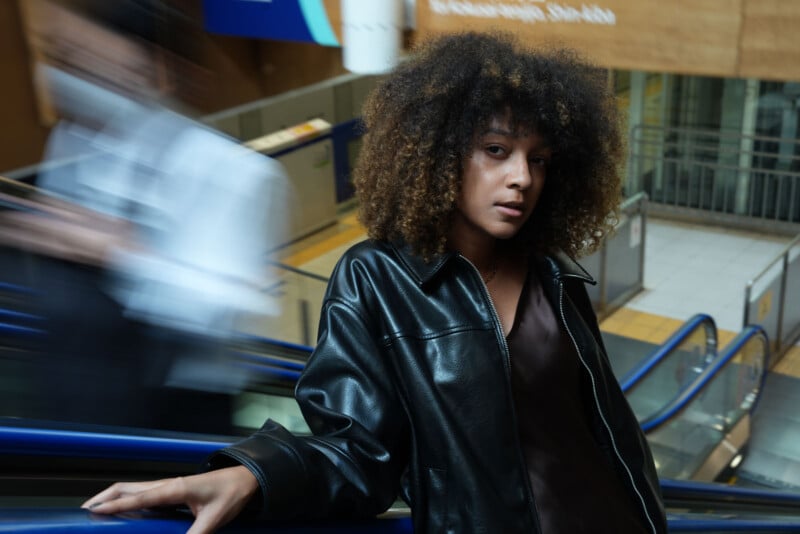 Una mujer con cabello rizado y una chaqueta de cuero negra se apoya en la barandilla de la escalera mecánica, mirando a la cámara mientras una figura borrosa pasa al fondo, creando una sensación de movimiento.