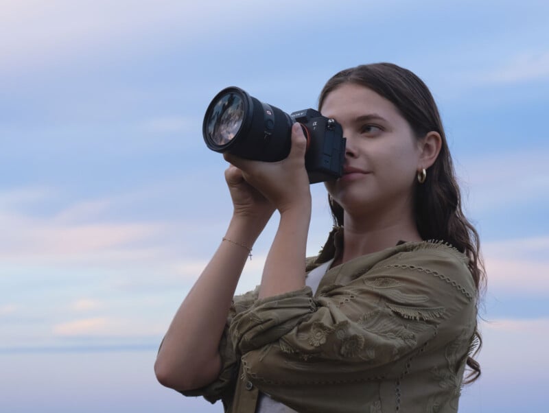 Una mujer joven con cabello largo y castaño, aretes y una chaqueta verde oliva sostiene una cámara frente a sus ojos, preparándose para tomar una foto al aire libre bajo un cielo suave y pastel.