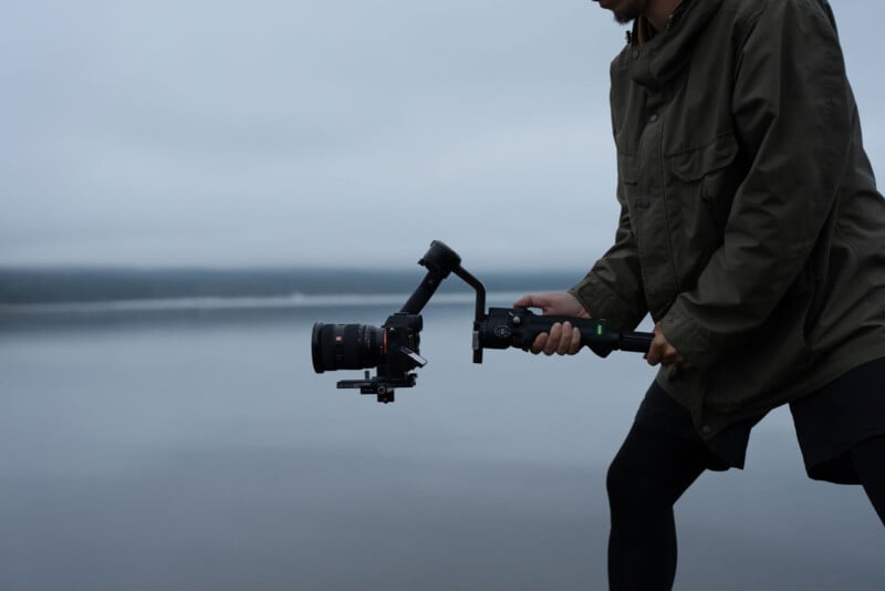 Un hombre con una chaqueta oscura maneja una cámara profesional montada en un estabilizador junto a una masa de agua tranquila y turbia. El fondo es borroso y gris, enfatizando el equipo de filmación.