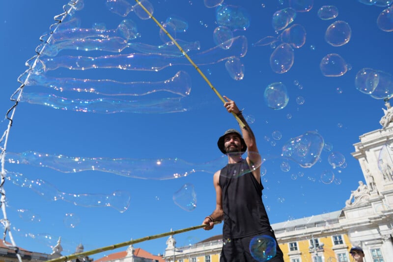 Un hombre con chaleco negro y sombrero de pescador hace grandes pompas de jabón con un palo y una cuerda en una soleada plaza de la ciudad, con el cielo azul y edificios históricos al fondo.