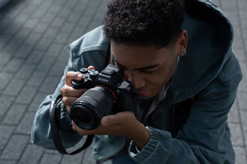 Un hombre con una chaqueta verde se agachó y tomó una fotografía a través del visor de una cámara Sony. El fondo muestra una superficie pavimentada con un patrón en forma de cuadrícula.