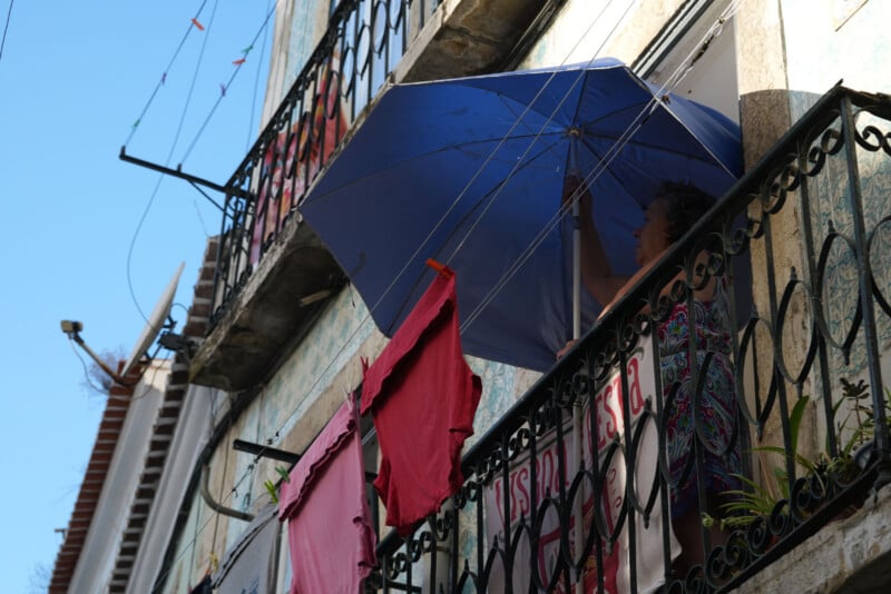 En el balcón había un hombre con un gran paraguas azul sobre la cabeza. Ropa colorida cuelga de las barandillas y el sol proyecta sombras sobre las paredes de azulejos del edificio. El cielo estaba claro y azul.
