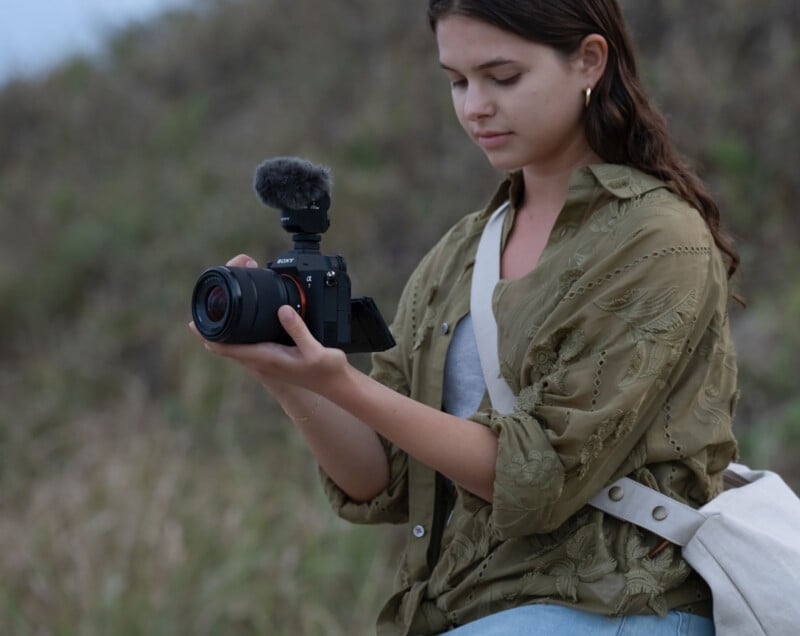 Una persona sostiene una cámara con un micrófono al aire libre y mira la pantalla. Llevaban camisas de color verde oliva y bolsas de colores claros colgadas de los hombros. El fondo está cubierto de hierba y borroso.
