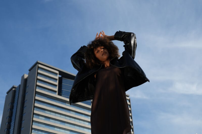 Una mujer de pelo rizado con un vestido negro y una chaqueta de cuero posa con confianza al aire libre con edificios modernos y un cielo azul de fondo.