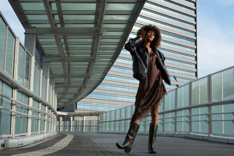 Una mujer de pelo rizado, vestida con una chaqueta de cuero negra, un vestido marrón y botas, se alza con confianza en un moderno puente peatonal con altos edificios de cristal al fondo.