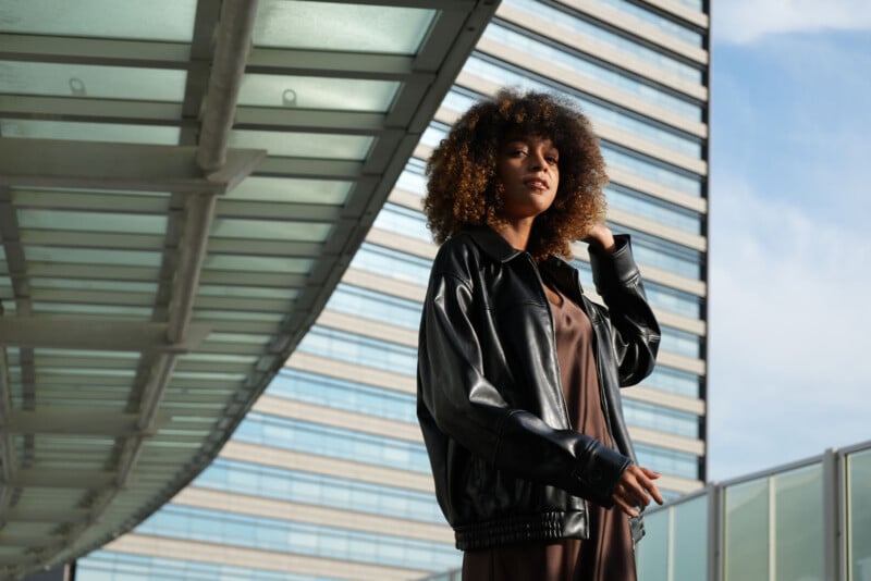Una mujer de cabello rizado, vestida con una chaqueta de cuero negra y un vestido marrón, se encuentra al aire libre bajo un moderno dosel de vidrio con un edificio alto y un cielo azul de fondo.