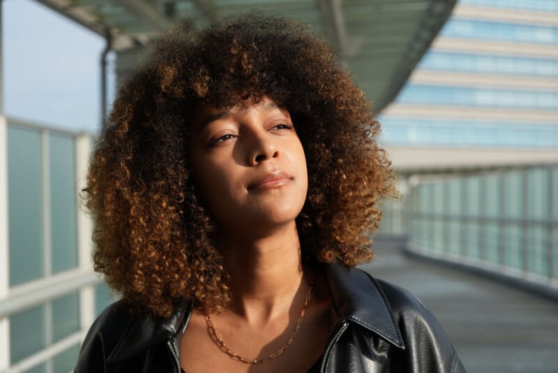 Una mujer joven con cabello rizado estaba parada bajo un dosel de vidrio al aire libre, vestida con una chaqueta de cuero negra y un collar de cadena de oro, mirando pensativamente a lo lejos con el sol brillando en su rostro.