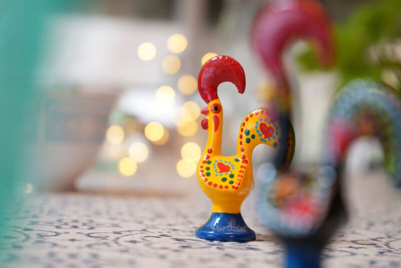 Una colorida estatuilla de cerámica de un gallo con patrones rojos, amarillos, azules y verdes se encuentra sobre una superficie estampada, con otro gallo borroso en primer plano y una iluminación suave al fondo.