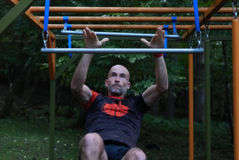 Un hombre con la cabeza afeitada y barba alcanza una barra en una carrera de obstáculos al aire libre, rodeada de árboles y espacios verdes.