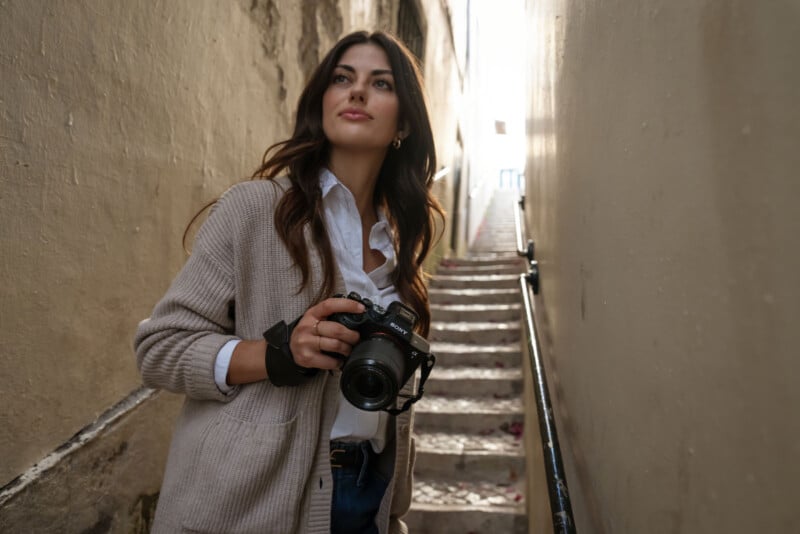 Una mujer de largo cabello castaño estaba parada en un callejón estrecho, sosteniendo una cámara en la mano y mirando hacia un lado. Detrás de ella, unos escalones de piedra conducen a la brillante luz del día. Llevaba una rebeca beige y una camisa blanca.