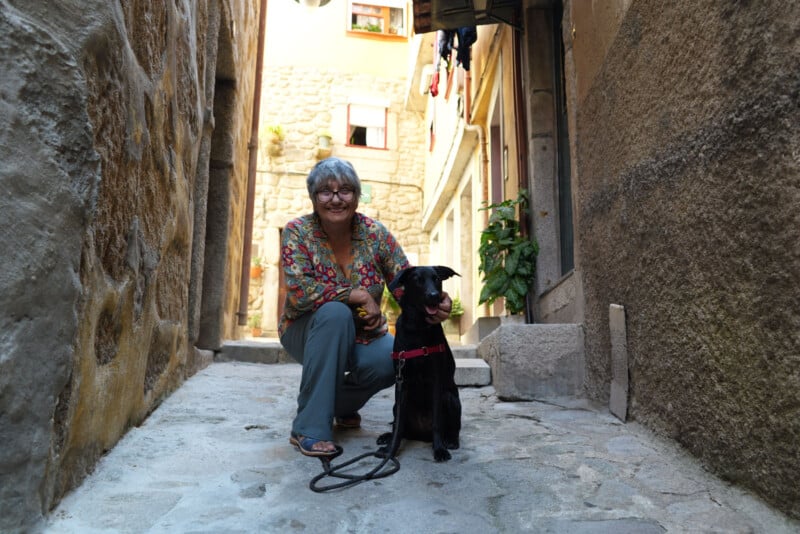 Una anciana se agacha junto a un perro negro con una correa y sonríe en un estrecho y soleado callejón de piedra bordeado de edificios y plantas antiguos.