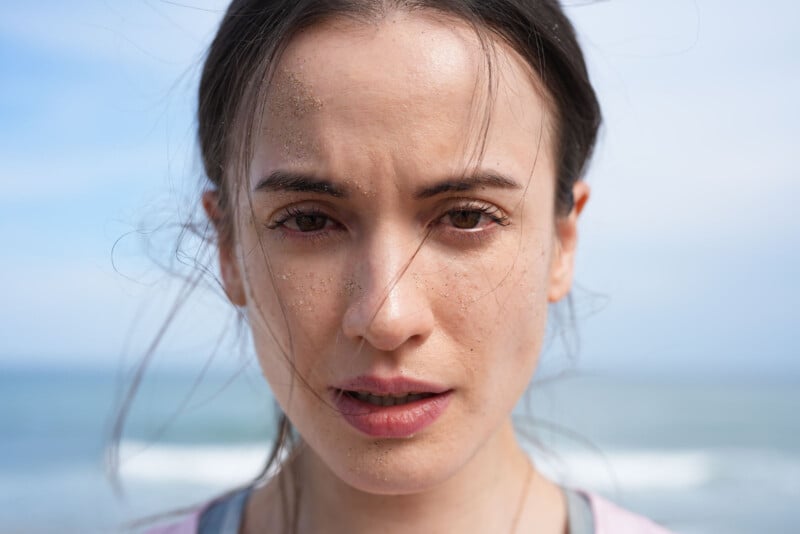 Una mujer joven con el rostro cubierto de arena mira directamente a la cámara. Mechones de su cabello castaño ondeaban al viento. El fondo borroso muestra el océano y el cielo azul.
