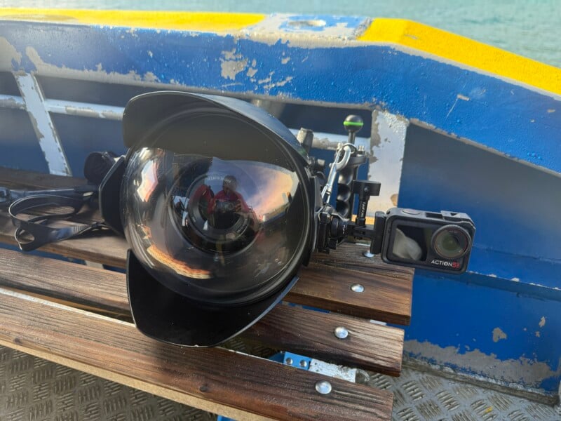 Hay una gran carcasa para cámara submarina con un puerto de cúpula y una cámara de acción sobre un banco de madera en un barco azul, con agua visible al fondo.