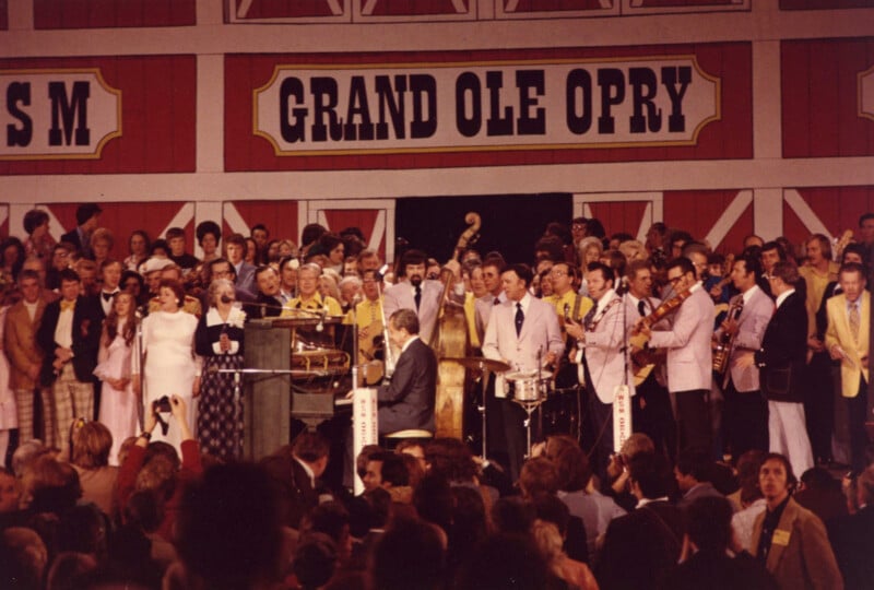 Un gran grupo de músicos e intérpretes se encontraba en el escenario del Grand Ole Opry mientras el público observaba. 