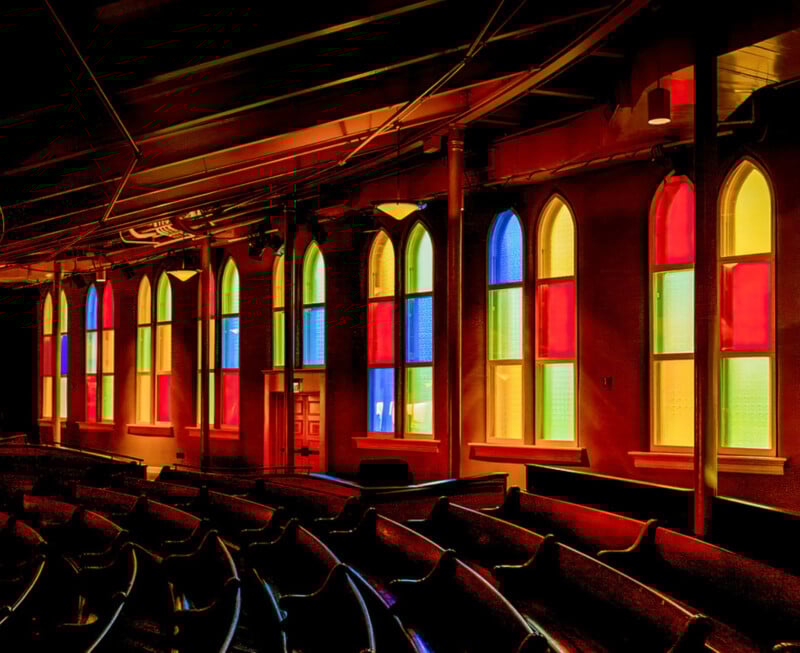 Una luz colorida brilla a través de grandes vidrieras arqueadas con paneles rojos, amarillos, azules y verdes, iluminando bancos de madera vacíos en iglesias o teatros con poca luz.