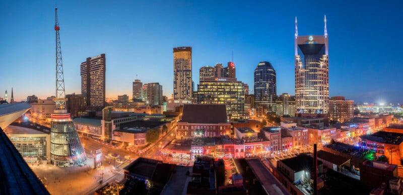 Vista panorámica del centro de Nashville al anochecer, incluidos edificios iluminados, la icónica Torre AT&T, luces de colores en calles concurridas y un cielo nocturno azul claro.