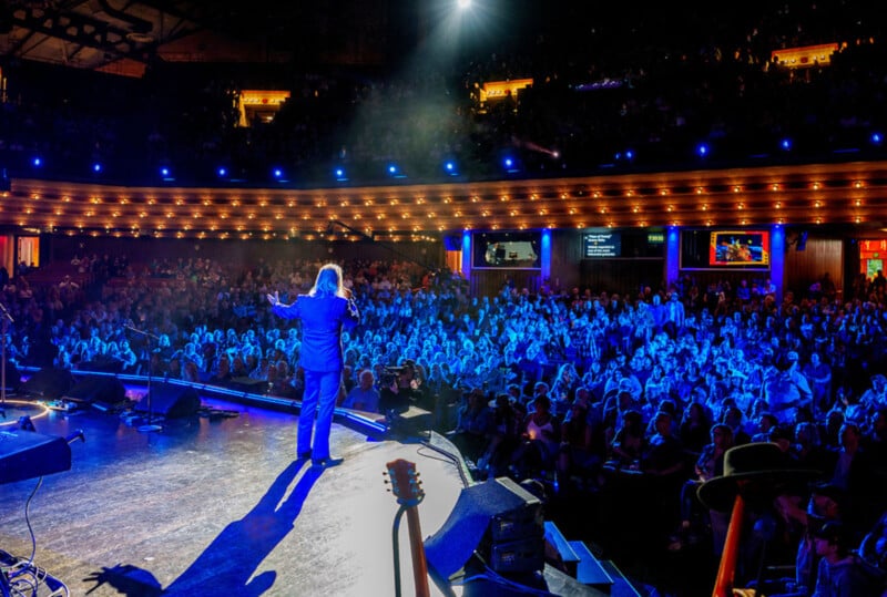 En un teatro, un artista se encuentra en un escenario brillantemente iluminado frente a una gran audiencia, luces azules iluminan a la multitud y cálidas luces amarillas decoran el balcón de arriba.