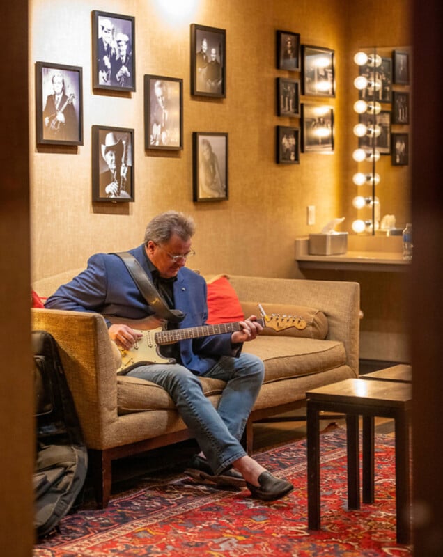 Un hombre con chaqueta azul y vaqueros está sentado en un sofá tocando una guitarra eléctrica en una habitación decorada con fotografías enmarcadas en blanco y negro e iluminada por luces.