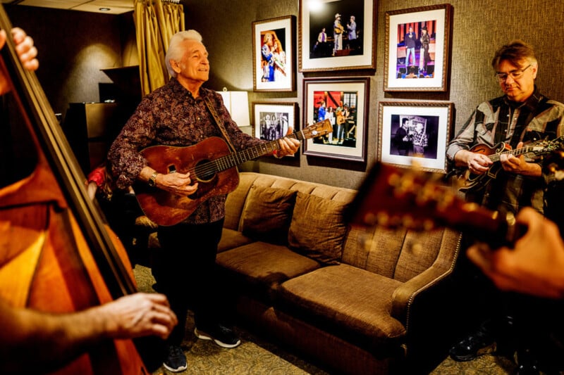 Un grupo de músicos tocaba la guitarra y la mandolina en una acogedora sala con marcos de cuadros en las paredes, mientras un hombre mayor estaba en el centro sonriendo y tocando una guitarra acústica.